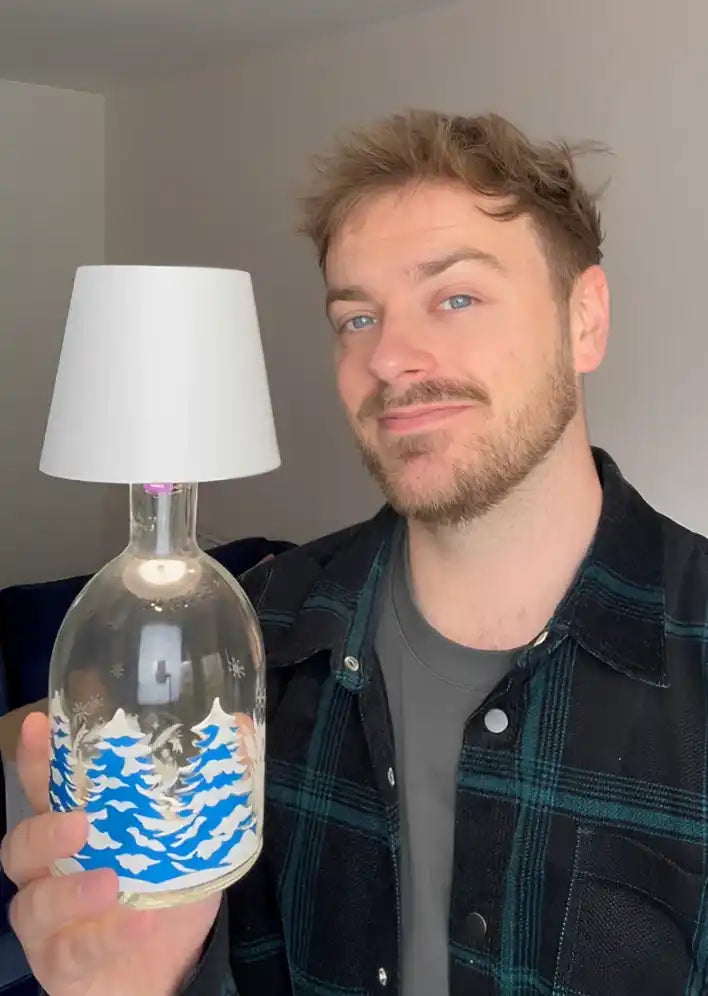 A clear glass bottle converted into a lamp with a white lampshade and blue and white winter landscape design.