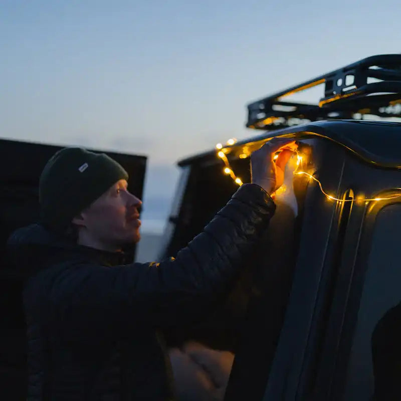 A person setting up string lights on a vehicle’s roof rack.