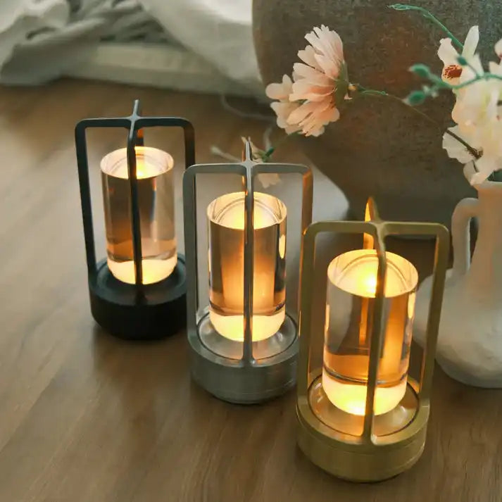 A trio of decorative lanterns, each featuring a clear glass cylinder with an illuminated amber glow, encased in distinct black, silver, and gold metal frames.